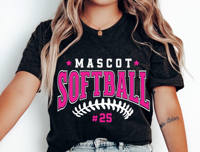 May include: A dark charcoal gray short-sleeve t-shirt featuring the text MASCOT SOFTBALL in bold pink letters with white outlines. Below the text is a white graphic of softball stitching and the pink text #25, flanked by two pink stars.