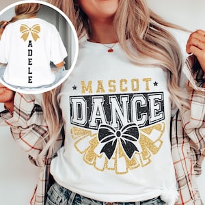 May include: A white t-shirt featuring a graphic print with the text MASCOT DANCE in black and gold glitter, accented by a black bow and gold pom-poms. An inset shows the back of the shirt with a gold glitter bow and the name ADELE in black text.