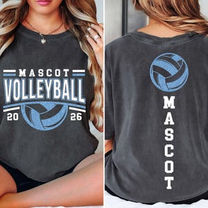 Op de afbeelding: Donkergrijs t-shirt met "MASCOT VOLLEYBALL" in wit en lichtblauw, een volleybalafbeelding en de jaren 20 en 26. De achterkant van het shirt heeft een volleybalafbeelding en het woord "MASCOT" verticaal.