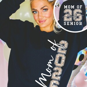 May include: Black sweatshirt with the text "Mom of 26" in a white script and "26" in a gold glitter font. The back of the sweatshirt has the text "Mom of 26 Senior" in white and gold glitter.