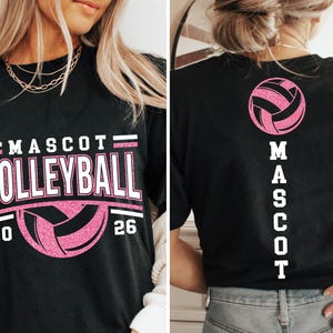Op de afbeelding: Zwart t-shirt met "MASCOT VOLLEYBALL 20 26" in witte en roze letters, met een volleybalafbeelding. De achterkant van het shirt toont een roze volleybal en het woord "MASCOT" verticaal.