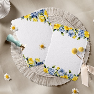 May include: Two blank white cards with a floral border of yellow and blue flowers. The cards are on a beige fringed mat with a small bouquet of dried yellow flowers.
