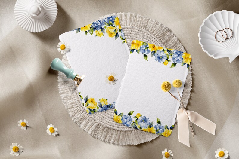 May include: Two blank white cards with a floral border of yellow and blue flowers. The cards are on a beige fringed mat with a small bouquet of dried yellow flowers.