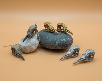 May include: A set of earrings featuring small bird skull designs. Two pairs of earrings are shown, one pair is gold and the other is silver. The earrings are displayed on a gray stone and a white rock.
