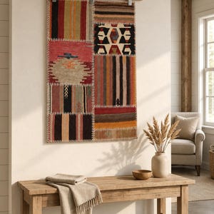 May include: A rectangular wall hanging made of stitched fabric panels featuring various geometric patterns and stripes in red, brown, black, and tan. The textile is suspended from a wooden rod with metal clips, displaying visible white stitching.