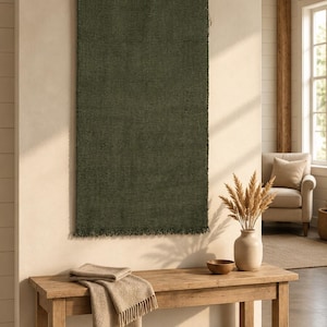 Peut inclure: Une tenture murale rectangulaire en tissu tissé vert olive avec un bord inférieur à franges, suspendue à une tringle en bois par trois pinces métalliques. Le textile présente un tissage dense et uniforme, affiché verticalement.