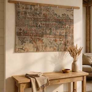 Op de afbeelding: Een rechthoekig geweven wandkleed met een vervaagd patroon in roestoranje, antracietgrijs en crème. Het textiel hangt aan een horizontale houten roede met vier metalen clips en vertoont een verweerd, vintage-geïnspireerd ontwerp.