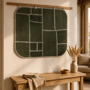May include: A square, dark olive green wall hanging with rounded corners and a grid-like pattern of light gray stitched lines. The textile piece is suspended from a horizontal wooden rod by four metal clips, displaying a textured surface.