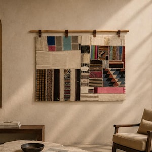 May include: A rectangular wall hanging tapestry composed of various fabric patches in diverse colors and patterns, including stripes, geometric shapes, and solid blocks, suspended from a horizontal wooden rod by four brown leather straps.
