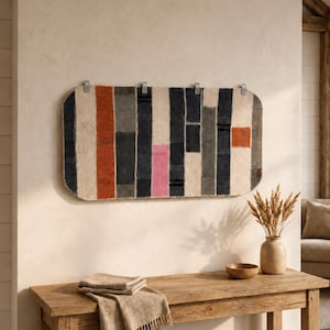 Puede incluir: Un tapiz rectangular con un patrón de rayas verticales en tonos crema, gris carbón, naranja quemado y un bloque rosa brillante. El tejido tiene esquinas redondeadas y está sujeto a la pared con cuatro clips de metal plateado.