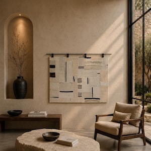 May include: A rectangular wall hanging featuring a patchwork of cream, beige, and white fabric panels with black linear and dashed patterns, suspended from a dark metal rod. The textile art displays a geometric composition of varied textures.