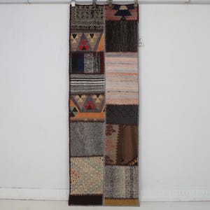 Könnte beinhalten: Ein rechteckiger Patchwork-Teppich aus verschiedenen gewebten Stoffsegmenten in unterschiedlichen Farben wie Orange, Braun, Grau und Schwarz. Der Teppich zeigt mehrere geometrische Muster, Streifen und einfarbige Texturen, die vertikal miteinander vernäht sind.