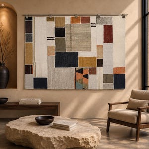 May include: A large rectangular wall tapestry featuring a geometric patchwork design composed of various fabric blocks in shades of cream, charcoal, mustard yellow, rust orange, olive green, and light grey, suspended from a metal rod.