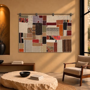 Puede incluir: Un tapiz de pared rectangular con un diseño de patchwork de varios segmentos de tela tejida en tonos rojo, naranja, marrón, gris, negro y crema, suspendido de una barra de metal oscuro con cuatro clips a juego sujetos al borde superior.
