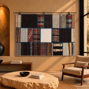 Op de afbeelding: Een rechthoekig wandtapijt met een patchworkontwerp van diverse stoffen rechthoeken in zwart, wit, grijs, rood en blauw, verbonden door zichtbare stiksels en opgehangen aan een horizontale metalen roede met drie clips.