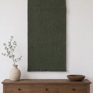 Puede incluir: Un tapiz rectangular de tejido color verde oliva oscuro con un borde inferior deshilachado, suspendido de una barra de madera horizontal mediante cuatro clips de metal plateado. La tela presenta un patrón de tejido denso y uniforme.