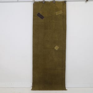 May include: A rectangular olive green textile wall hanging featuring a frayed bottom edge and three small, rectangular patches in dark brown, light beige, and tan. The fabric is suspended from a metal rod by four silver metal clips.
