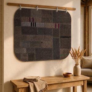May include: A rectangular dark gray textile wall hanging with rounded corners, featuring a patchwork design of various woven textures and small magenta and green accents, suspended from a horizontal wooden rod by four metal clips.