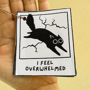 Può includere: Un'etichetta quadrata bianca con bordo nero che presenta l'illustrazione di un gatto nero che salta attraverso uno sfondo incrinato. Il testo I FEEL OVERWHELMED è stampato in nero in basso. È presente una catenella a palline in metallo.