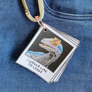 Peut inclure: Une pile de petites étiquettes carrées avec une image de dinosaure et le texte "I WOULD LIKE TO LEAVE". Le dinosaure est bleu et gris, avec un autocollant d'œil. Les étiquettes sont attachées à un mousqueton argenté.
