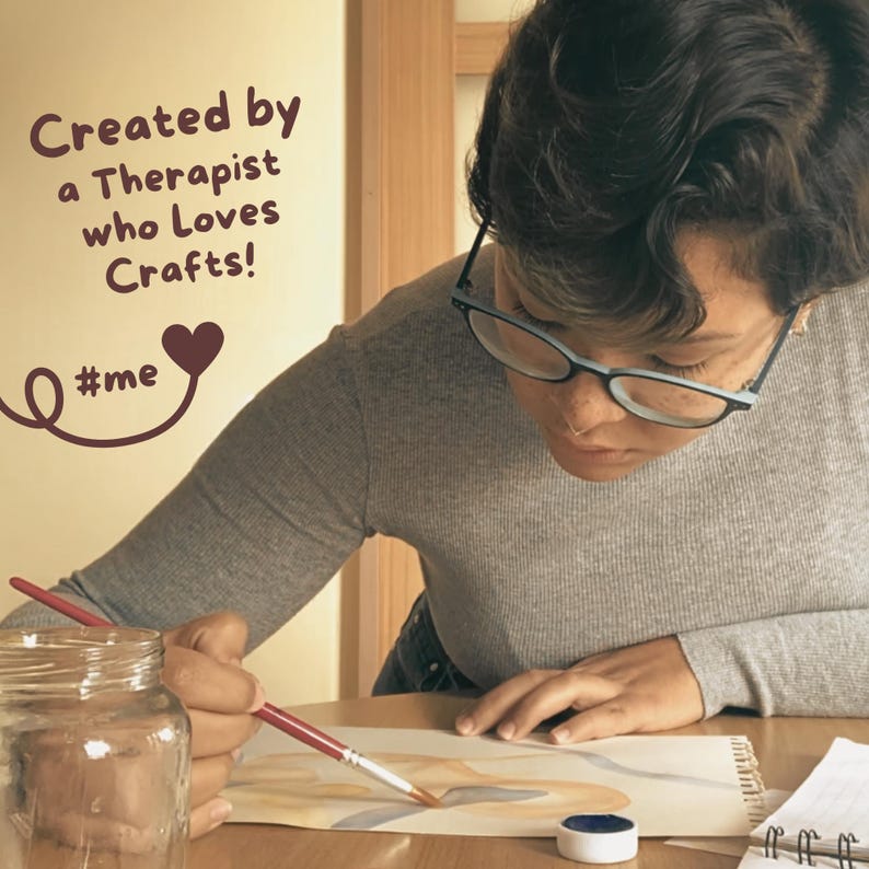 K&ouml;nnte beinhalten: Eine Person mit Brille malt mit einem Pinsel auf ein Blatt Papier. Das Papier liegt auf einem Holztisch. Auf dem Tisch stehen ein Glas und ein kleiner Beh&auml;lter mit Farbe. Der Text "Created by a Therapist who Loves Crafts! #me" ist an der Wand hinter der Person geschrieben.