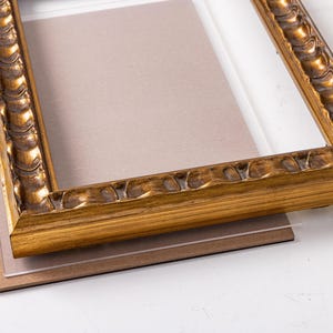 May include: A gold picture frame with a decorative, ornate design. The frame is made of wood and has a glass front. The frame is sitting on a brown mat.