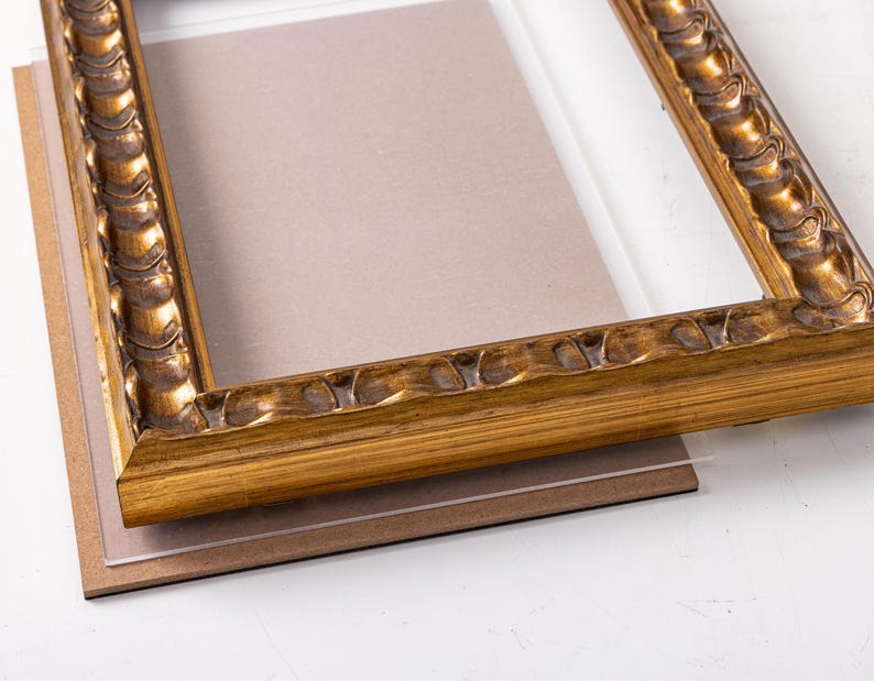 May include: A gold picture frame with a decorative, ornate design. The frame is made of wood and has a glass front. The frame is sitting on a brown mat.