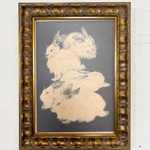 May include: A framed print of a black and white illustration of four white rabbits stacked on top of each other. The rabbits are depicted in a realistic style with detailed fur and whiskers. The print is framed in a gold-colored frame with a decorative border.