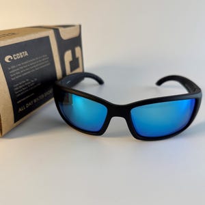 May include: Black sunglasses with reflective blue lenses and a matte black frame. The sunglasses are positioned in front of a brown cardboard box featuring the Costa logo and text that reads All Day Water Sport in black and white print.