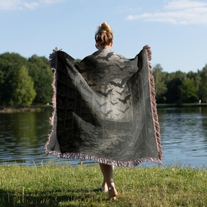 Puede incluir: Una manta gris oscuro con un dise&ntilde;o de murci&eacute;lagos y estanter&iacute;as. La manta tiene un borde con flecos y se sostiene frente a un lago. El dise&ntilde;o presenta murci&eacute;lagos volando frente a una estanter&iacute;a.