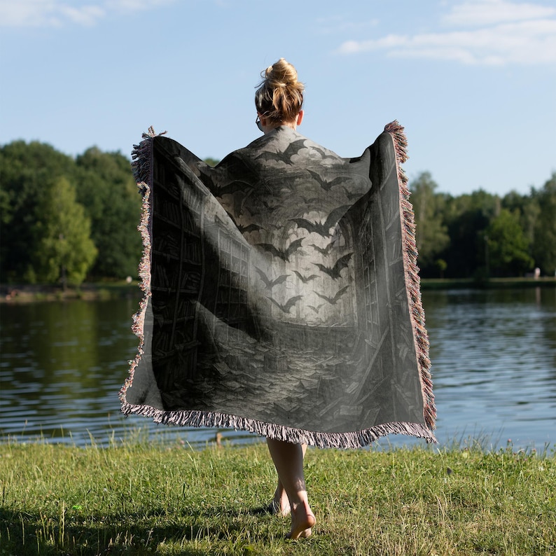Puede incluir: Una manta gris oscuro con un dise&ntilde;o de murci&eacute;lagos y estanter&iacute;as. La manta tiene un borde con flecos y se sostiene frente a un lago. El dise&ntilde;o presenta murci&eacute;lagos volando frente a una estanter&iacute;a.