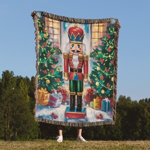 May include: A woven throw blanket featuring a colorful illustration of a nutcracker soldier. The design includes Christmas trees, wrapped gifts, and a window scene. The blanket has a fringed edge and is held up against a blue sky.