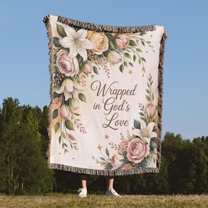 May include: A cream-colored throw blanket with a floral design featuring pink roses, white lilies, and peach-colored flowers. The blanket has a brown fringe and the text "Wrapped in God's Love".