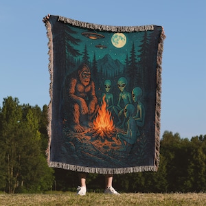 May include: A woven throw blanket featuring a campfire scene with a Bigfoot, aliens, and UFOs under a full moon. The blanket has a dark blue background with a fringed edge.