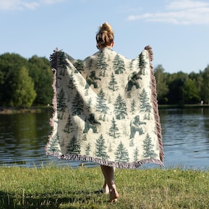 May include: A cream-colored throw blanket featuring a pattern of green pine trees and Bigfoot figures. The blanket has a fringed edge with brown and purple threads. The person holding the blanket is standing near a lake.