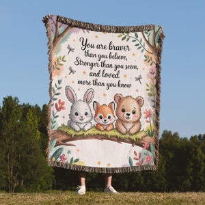 May include: A woven throw blanket featuring a whimsical design with a rabbit, fox, and bear on a branch. The blanket has the text "You are braver than you believe, stronger than you seem, and loved more than you know."