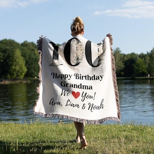 Peut inclure: Une couverture tissée blanche avec du texte noir qui dit "100 Joyeux anniversaire Grand-mère Nous ♥ Vous! Ava, Liam & Noah". La couverture a un motif floral et est tenue par une personne dans un parc.