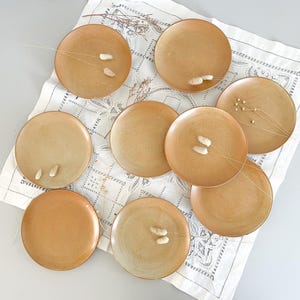 Op de afbeelding: Een verzameling van negen ronde, bruin-beige keramische borden, gerangschikt op een witte, bedrukte doek. Elk bord heeft een iets andere bruin-beige tint. Sommige borden zijn versierd met kleine, witte, langwerpige accenten en gedroogde sprietjes.