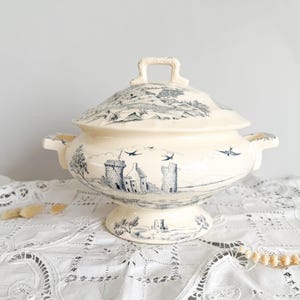 May include: Cream ceramic tureen with a lid, decorated with a blue and white landscape scene. The tureen has a pedestal base, handles, and a square handle on the lid. The design includes castles, trees, and birds in flight.