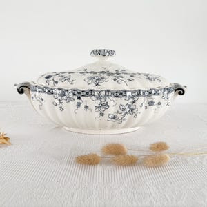May include: A white and blue floral patterned serving dish with a lid. The dish has two handles and a decorative rim. The lid is topped with a small knob. The dish is on a white surface.