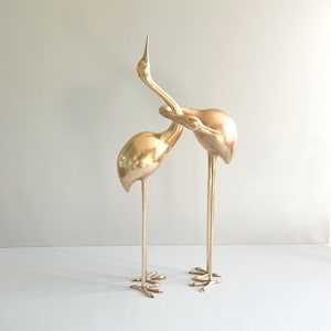 May include: Two gold-colored crane sculptures. The cranes have long, slender legs and necks, with one crane's neck intertwined with the other. The sculptures are made of a shiny, metallic material and stand on a white surface.
