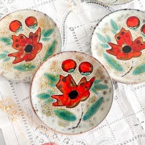 May include: A set of four decorative ceramic plates. Each plate features a red poppy flower with a black center, red berries, and green leaves on a cream-colored background. The plates have a textured, crackled glaze.
