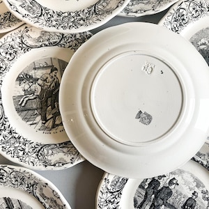 K&ouml;nnte beinhalten: Ein Set antiker wei&szlig;er Keramikteller mit Schwarzwei&szlig;illustrationen. Die Teller zeigen Szenen von Menschen, die in einer Fabrik arbeiten. Die R&uuml;ckseite eines Tellers tr&auml;gt einen Stempel mit der Aufschrift "Pour la memoire".
