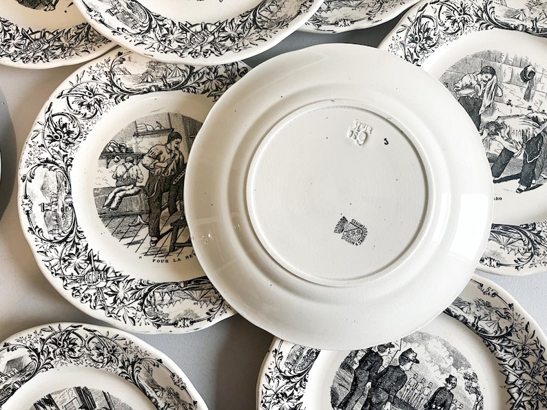 K&ouml;nnte beinhalten: Ein Set antiker wei&szlig;er Keramikteller mit Schwarzwei&szlig;illustrationen. Die Teller zeigen Szenen von Menschen, die in einer Fabrik arbeiten. Die R&uuml;ckseite eines Tellers tr&auml;gt einen Stempel mit der Aufschrift "Pour la memoire".