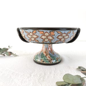 Peut inclure: Un bol sur pied en céramique avec un bord et une base brun foncé. L'extérieur du bol présente un motif répétitif de fleurs bleu clair, beige et blanc. Le pied est décoré d'un design marbré dans des tons de vert, de bleu et de brun.
