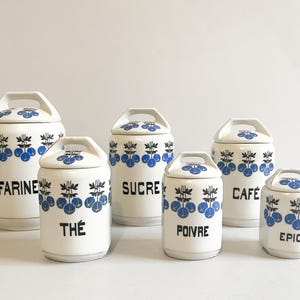 May include: Set of six white ceramic canisters with blue and black floral designs. The canisters are labeled "Farine", "The", "Sucre", "Poivre", "Cafe", and "Epices".