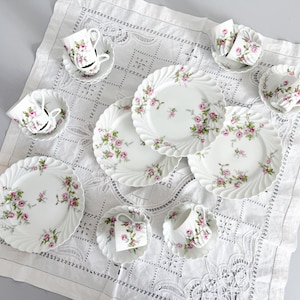 May include: A set of white porcelain teacups, saucers, and plates decorated with pink roses. The plates have a scalloped edge. The items are arranged on a white, patterned tablecloth.