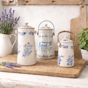 May include: A collection of ceramic canisters and a pitcher with a farmhouse aesthetic. The canisters are off-white with blue floral patterns and cow illustrations. A wooden cutting board and a white pitcher with lavender complete the scene.