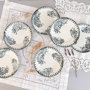 Puede incluir: Un juego de seis platos de cerámica blanca con un diseño floral verde oscuro. Los platos tienen un borde festoneado y están colocados sobre un mantel blanco estampado. Flores secas están esparcidas alrededor de los platos.