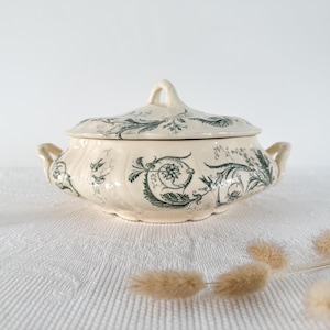 Puede incluir: Fuente de servir crema con tapa, con un patrón floral y de hojas en verde oscuro. La fuente tiene dos asas y un asa en la tapa. La fuente está sobre una superficie blanca.